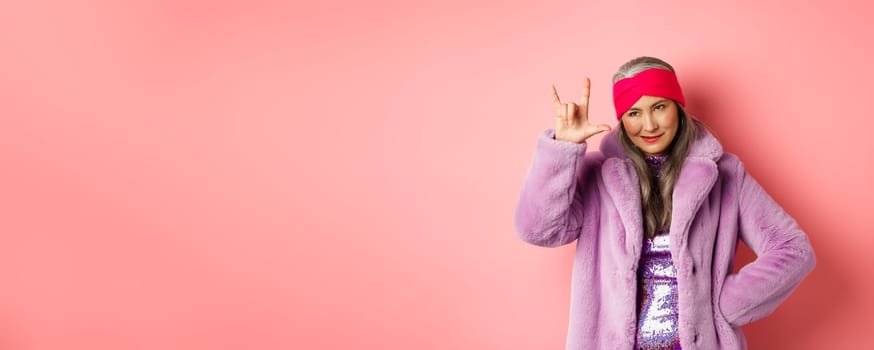 Funny and cool asian senior woman showing rock n roll gesture, looking sassy, standing over pink background