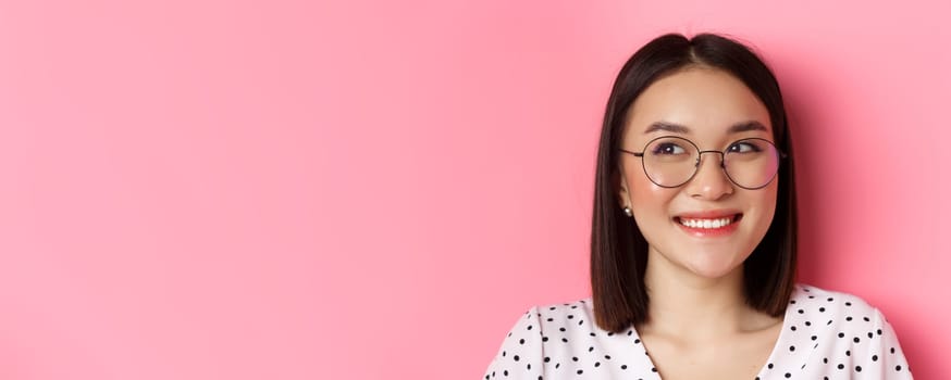 Beauty and lifestyle concept. Close-up of cute asian female model wearing trendy glasses, smiling and looking left at copy space, standing on pink background