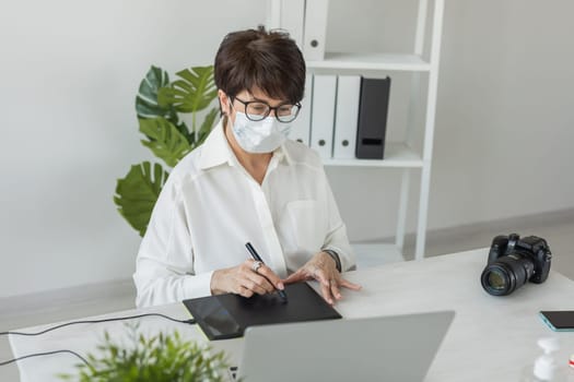Mature intelligent woman with medical mask working with computer and graphic tablet and stylus. She is a successful self employed retoucher and photograph
