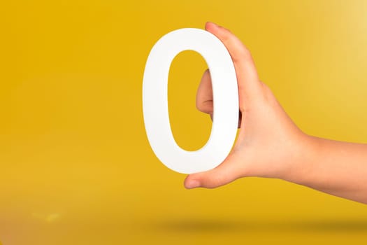 Numeral zero in hand. A hand holds a white number zero on a yellow background with copy space. Zero concept, 0 percent interest rate, minimum air emissions, cost or credit no increase.