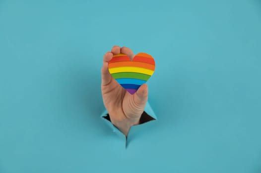 A hand with a rainbow-colored heart sticking out of a hole in a blue cardboard background.