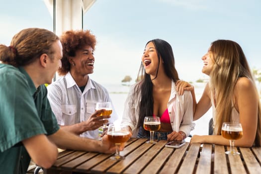 Group of multiracial young friends laughing, talking and having fun at beach bar drinking beers together.