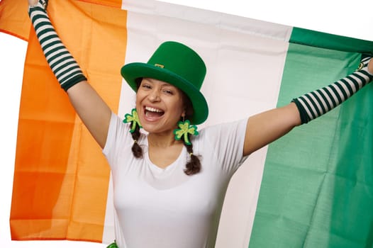 Cheerful ethnic woman wearing green St Patrick's hat and clover leaves earrings, carrying Irish flag on white background