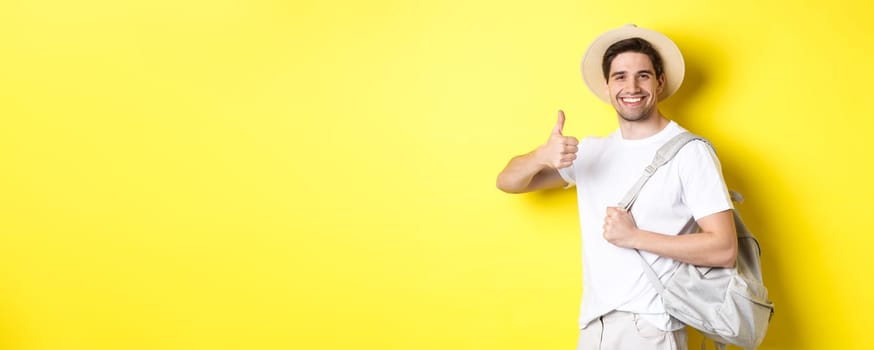 Tourism, travelling and holidays concept. Happy man going on vacation, holding backpack and showing thumb up, smiling satisfied, standing against yellow background