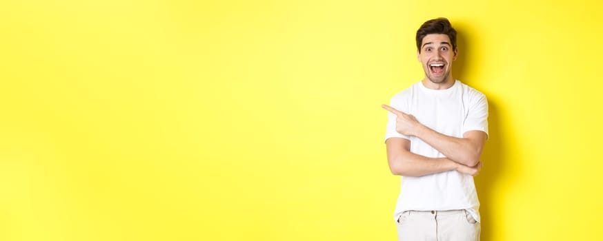 Happy man pointing finger left, showing advertisement on copy space, smiling amused, standing in white clothes against yellow background