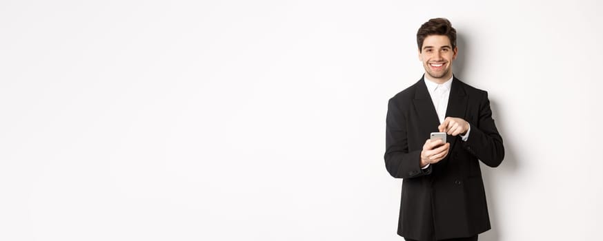 Image of handsome businessman in suit showing you app on smartphone, pointing finger at mobile screen and smiling, standing over white background