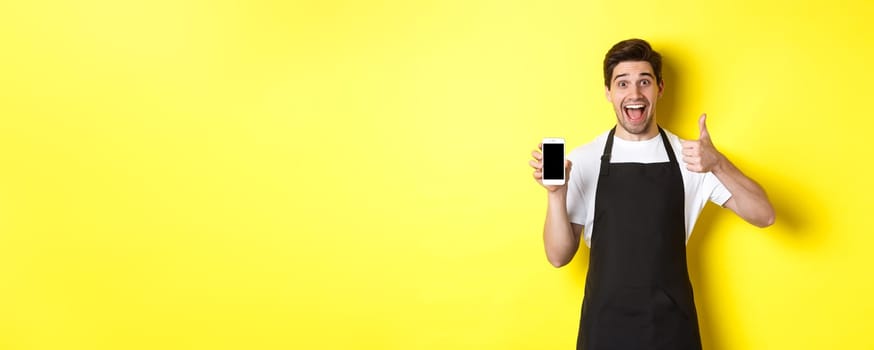 Happy barista in black apron showing smartphone screen, make thumb-up, recommending cafe application, standing over yellow background