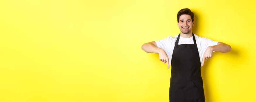 Friendly barista in black apron pointing fingers down, showing your logo banner, standing over yellow background