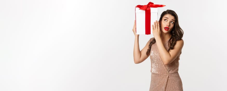 Holidays, celebration concept. Beautiful woman in luxury dress trying to guess what inside gift box, shaking Christmas present with curious face, white background.