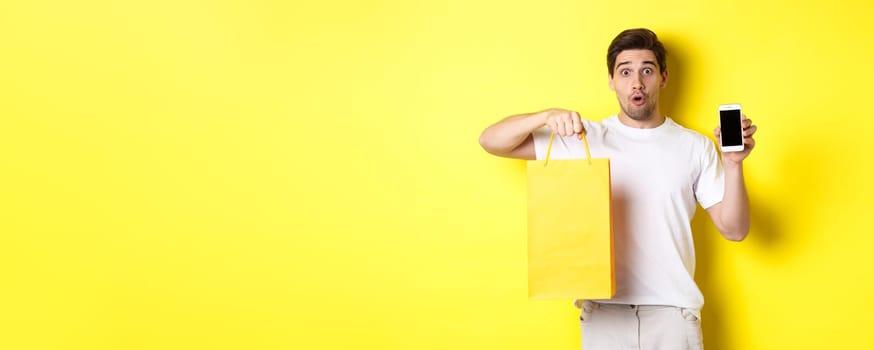 Surprised man holding shopping bag and showing smartphone screen, concept of mobile banking and app achievements, yellow background