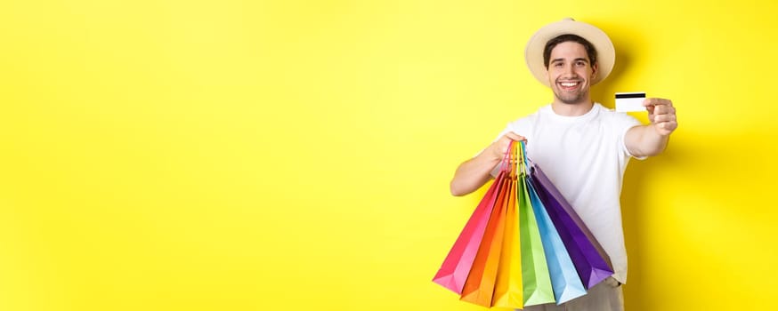 Smiling young man buying things with credit card, holding shopping bags and looking happy, standing over yellow background