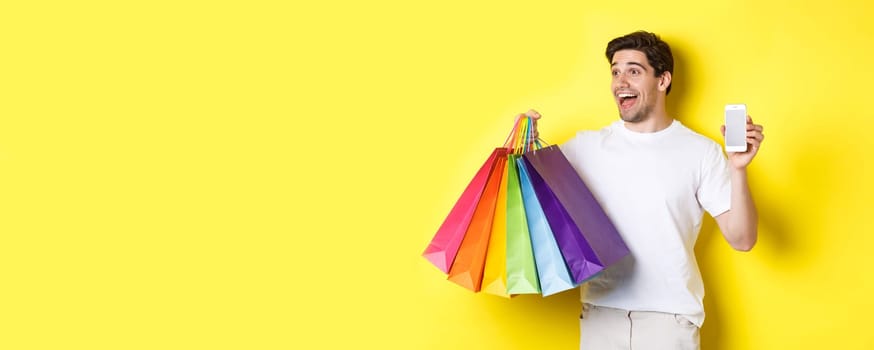 Excited man showing smartphone screen and shopping bags, achieve app goal, demonstrating mobile banking application, yellow background