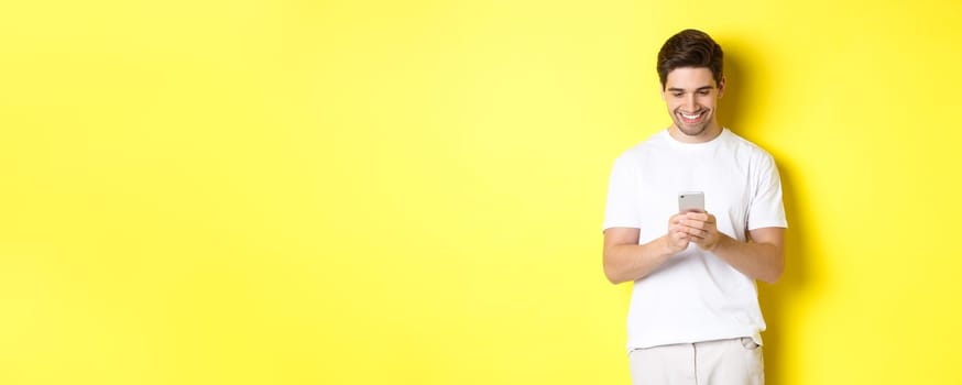 Young man reading text message on smartphone, looking at mobile phone screen and smiling, standing in white t-shirt against yellow background