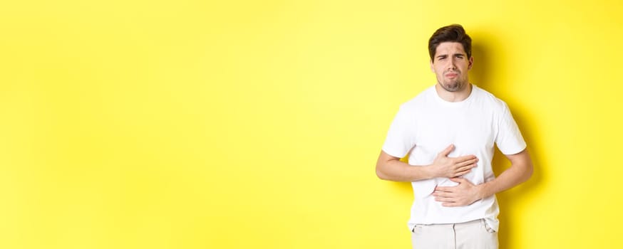 Man having stomach ache, grimacing from pain and touching belly, standing over yellow background