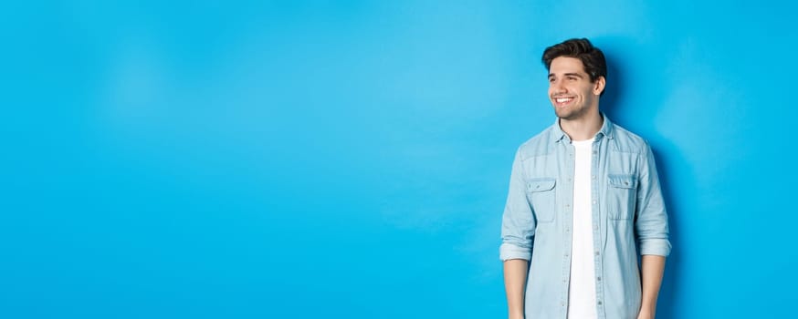 Handsome smiling adult man in casual outfit, smiling and looking left at promo offer, standing against blue background