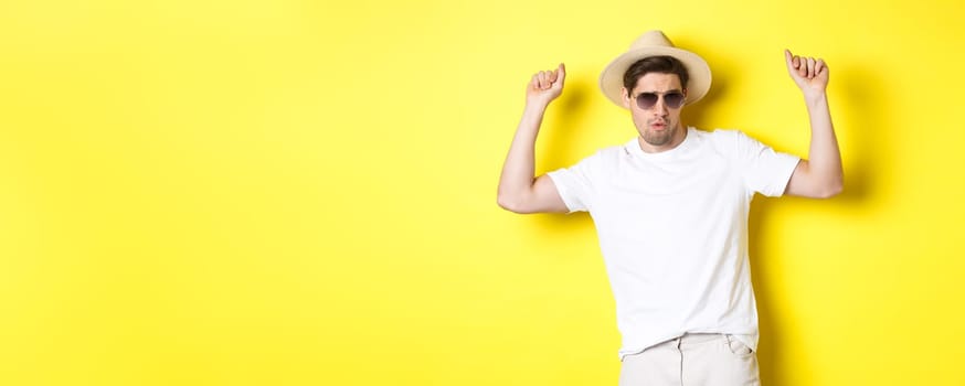 Tourism, travelling and holidays concept. Man tourist enjoying vacation, dancing in straw hat and sunglasses, posing against yellow background