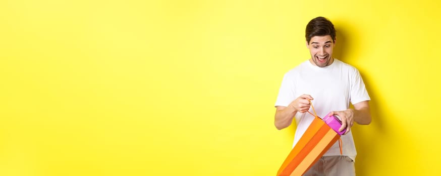 Concept of holidays and celebration. Young man looking surprised as take out gift from shopping bag, standing over yellow background