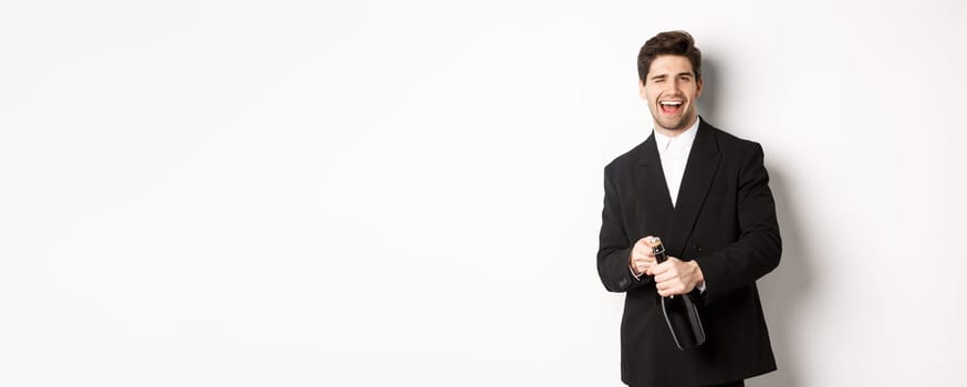 Image of handsome, confident man in black suit, celebrating holiday, open a bottle of champagne and partying, standing joyful against white background