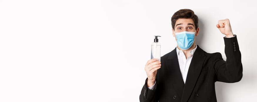 Concept of covid-19, business and social distancing. Close-up of happy man in trendy suit and medical mask, cheering and raising hand up, showing hand sanitizer, white background
