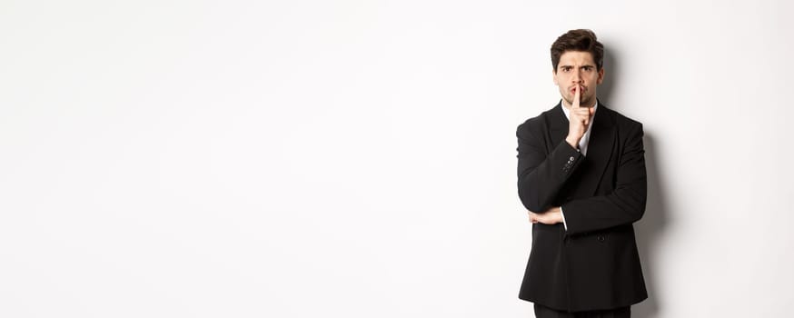 Portrait of angry boss in suit shushing at you, telling to be quiet, showing taboo hush sign and frowning, standing over white background