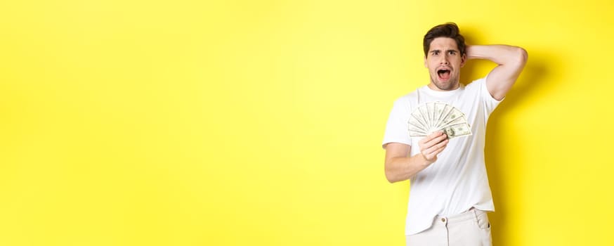 Frustrated man holding money, shouting and panicking, standing over yellow background