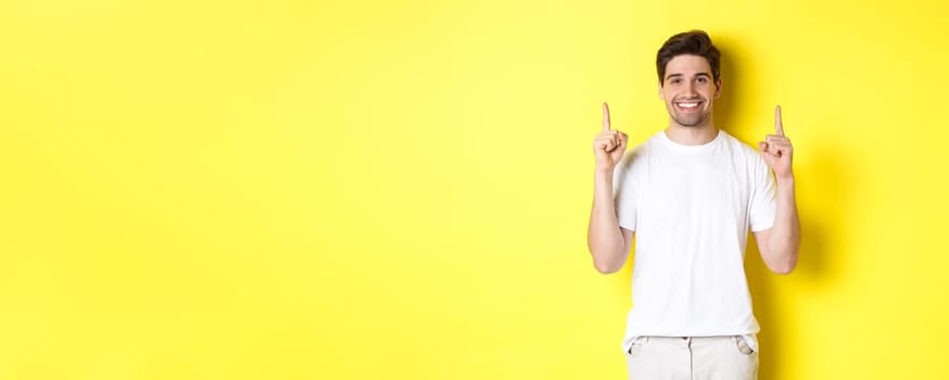 Handsome guy in white t-shirt pointing fingers up, showing shopping offers, standing over yellow background