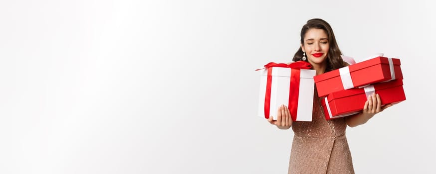 Holidays, celebration concept. Beautiful caucasian woman in elegant dress holding Christmas presents and smiling happy, standing over white background