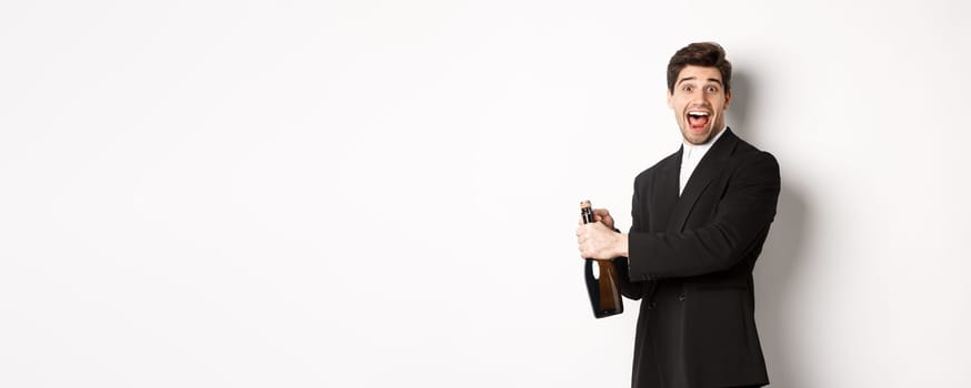 Portrait of attractive man in black suit, winking at camera and opening bottle of champagne, celebrating new year, standing against white background