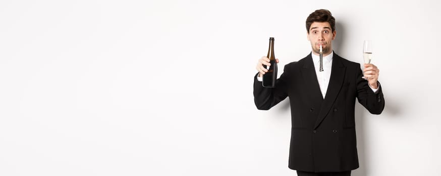 Concept of holidays, party and celebration. Portrait of handsome guy in black suit, raising bottle of champagne and glass, blowing a party whistle, having a birthday, standing over white background