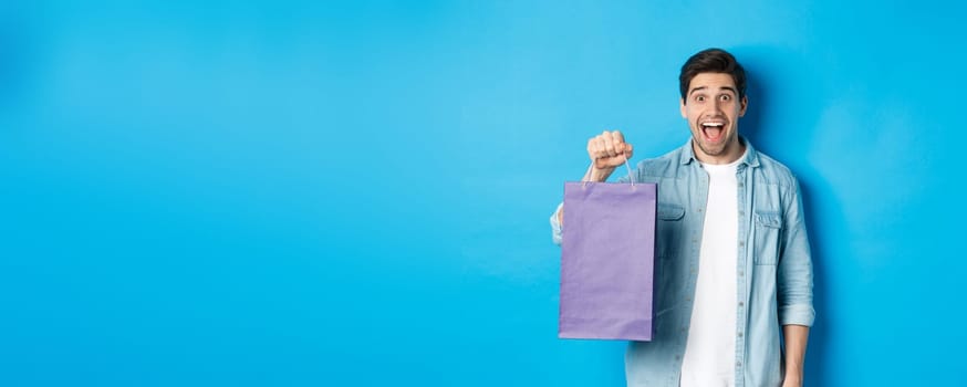 Concept of shopping, holidays and lifestyle. Excited handsome guy holding paper bag with gift and looking happy, standing over blue background