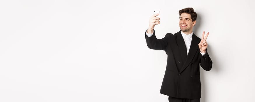 Portrait of good-looking man taking selfie on new year party, wearing suit, taking photo on smartphone and showing peace sign, standing against white background