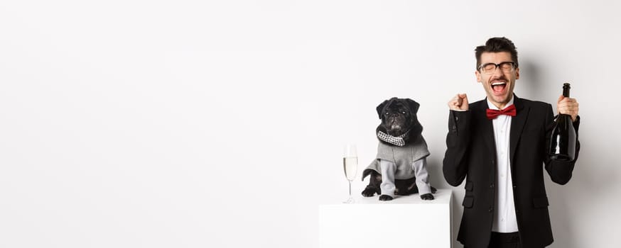 Pets, winter holidays and New Year concept. Happy young man celebrating Christmas with cute black dog wearing party costume, holding bottle champagne, white background