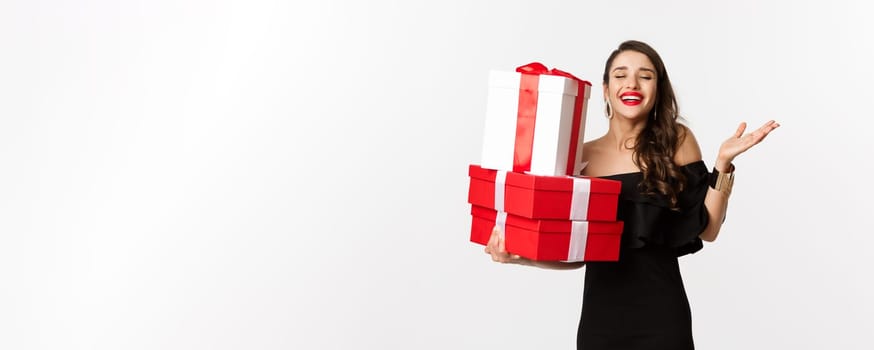 Celebration and christmas holidays concept. Excited and happy woman receive gifts, holding xmas presents and rejoicing, standing in black dress over white background
