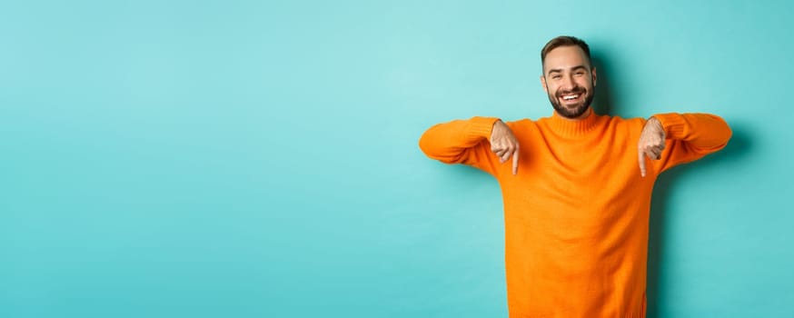Happy adult man with beard pointing fingers down, smiling cheerful, showing advertisement, standing over turquoise background