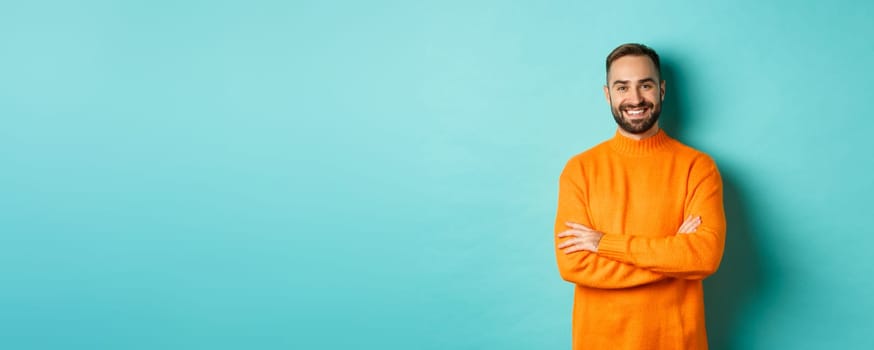 Handsome bearded male model in orange sweater, looking confident and smiling, cross arms on chest, standing over turquoise background