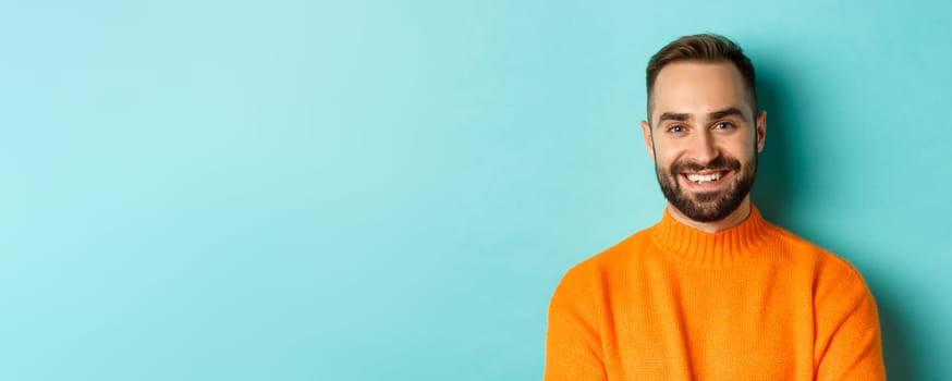 Close-up of handsome caucasian man smiling at camera, looking confident, wearing orange sweater, standing against turquoise background