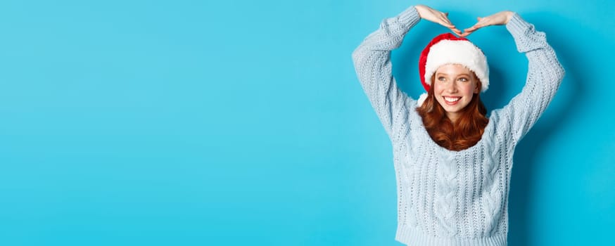 Winter holidays and Christmas Eve concept. Cute redhead teen girl in santa hat and sweater, making heart sign and smiling, wishing merry xmas, standing over blue background