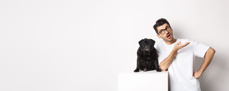 Image of hipster guy talking with his dog, looking skeptical and gesturing, having conversation with cute black pug, standing over white background
