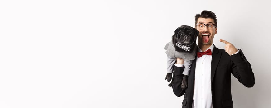 Funny young man in party suit, showing tongue and making grimaces, pointing at cute black dog in winter clothes, standing over white background