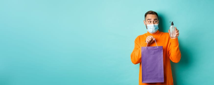 Coronavirus, pandemic and lifestyle concept. Man in face mask showing shopping bag and hand sanitizer, standing over turquoise background