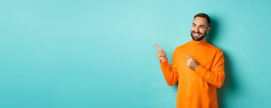 Attractive bearded male model in orange sweater, smiling satisfied and pointing fingers left, standing over turquoise background