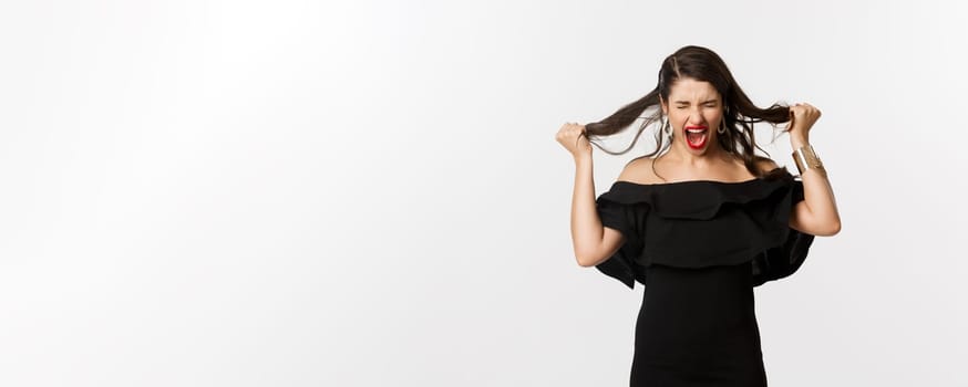 Fashion and beauty. Young woman in black dress screaming and rip hair on head, screaming mad, standing angry and outraged over white background.