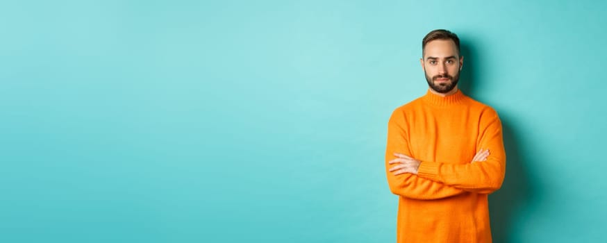 Confident young man looking determined, cross arms on chest, wearing orange winter sweater, standing against turquoise studio background