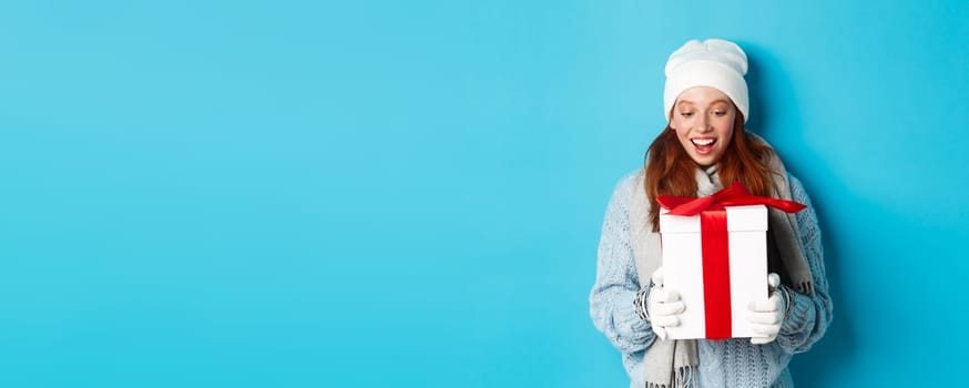 Winter holidays and Christmas eve concept. Surprised cute redhead girl in beanie and sweater receiving New Year gift, looking at present amazed, standing over blue background