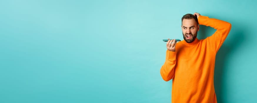 Confused man scratching head while talking on speakerphone, record voice message with indecisive face, standing in orange sweater over light blue background