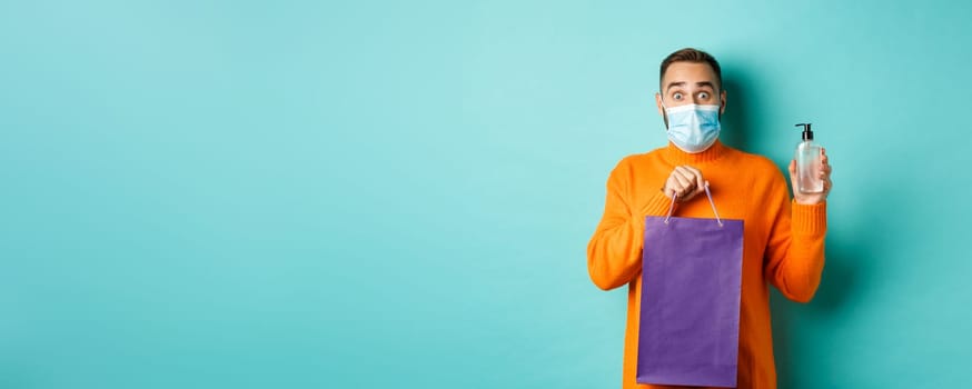Coronavirus, pandemic and lifestyle concept. Man in face mask showing shopping bag and hand sanitizer, standing over turquoise background.