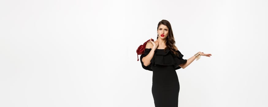 Beauty and fashion concept. Full length of arrogant and sassy woman in black dress, holding purse on shoulder and stroll forward at camera, posing over white background.