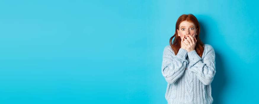 Shocked redhead girl in sweater, staring at camera scared and gasping, covering mouth with hands, standing against blue background