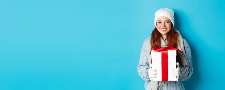 Winter holidays and Christmas sales concept. Cute redhead teeanage girl in beanie, sewater and scarf holding gift in wrapped box, smiling, wishing merry xmas, standing over blue background