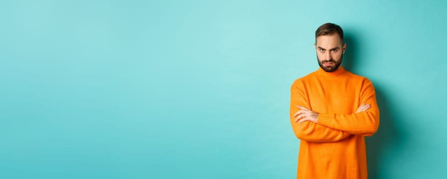 Offended man looking angry at you, cross arms on chest and stare mad, standing in orange sweater against turquoise background